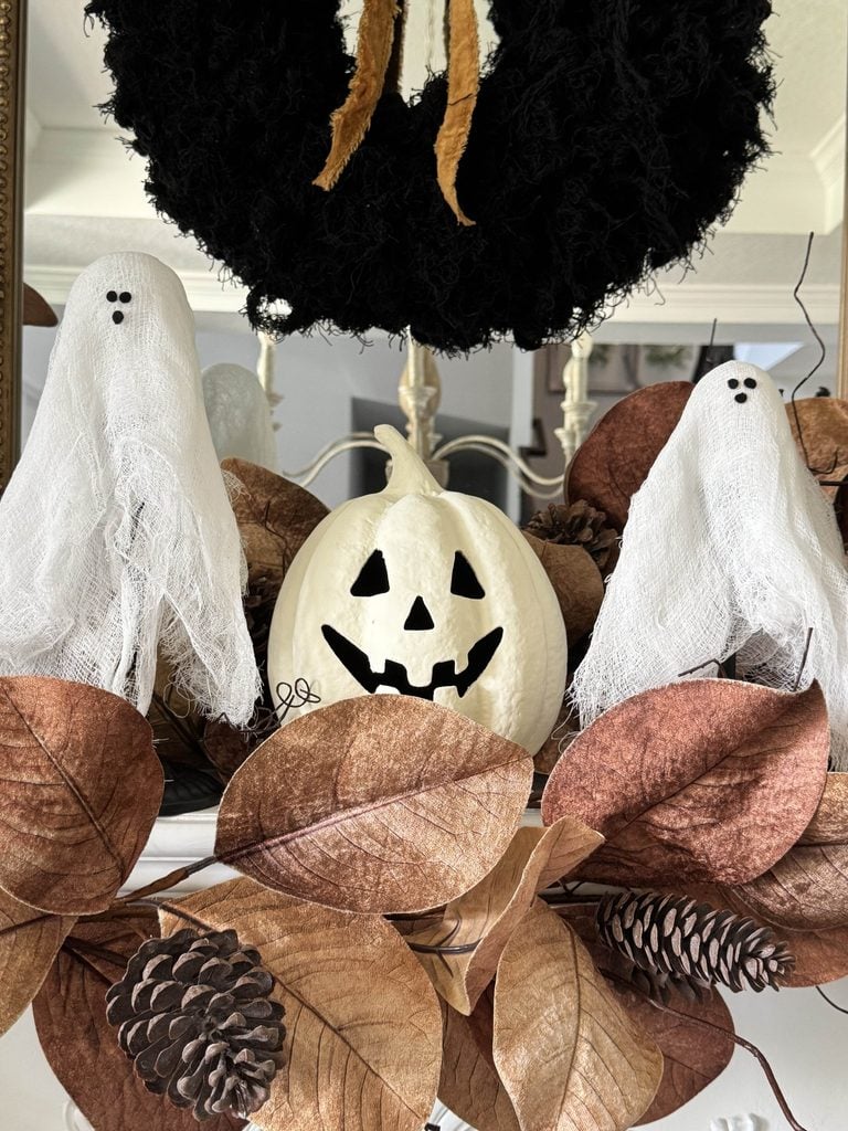 Two small ghost decorations and a white jack-o-lantern sit among brown leaves and pinecones, with a black wreath above—a charming Halloween mantel decor display.