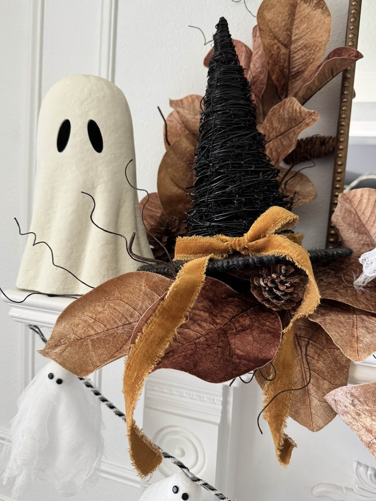 A white ghost figurine and a black witch hat with brown foliage, pinecone, and a gold ribbon above small hanging ghost ornaments.