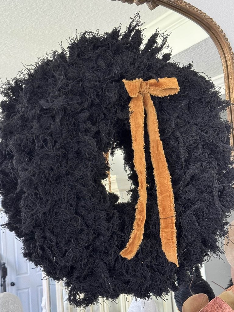 A fluffy black wreath with a soft, orange ribbon bow hangs on a mirror, creating stylish Halloween mantel decor. The reflection reveals part of a room with white walls and a door in the background.
