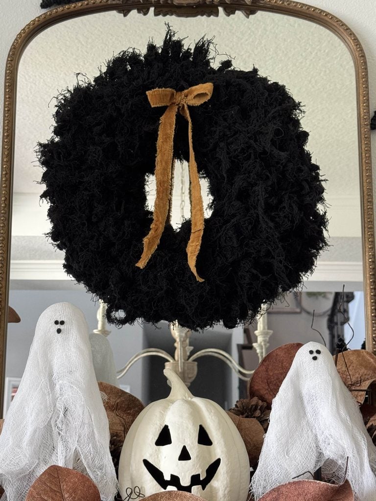 A black wreath with a gold bow hangs on a mirror. Below it, two small white ghost figures and a white jack-o-lantern rest among brown leaves.