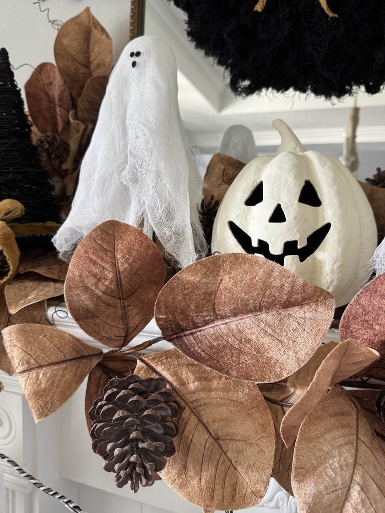 A white ghost decoration and a jack-o-lantern pumpkin are surrounded by brown leaves and a pinecone, making charming Halloween mantel decor for an autumn-themed display.