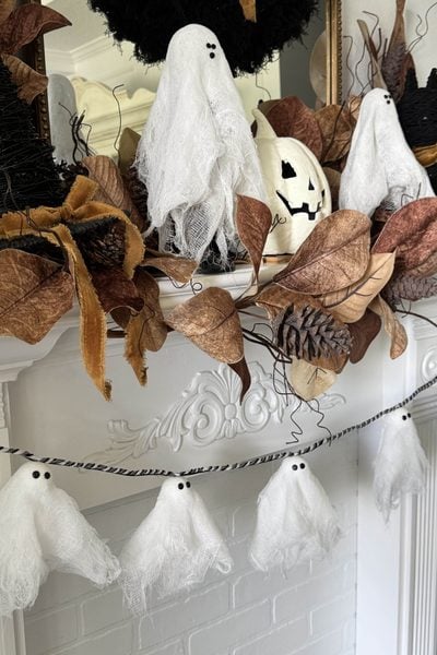 White gauze ghosts and brown leaves decorate a white mantel. A jack-o-lantern, black witch hats, pinecones, and a garland with mini ghosts hang below, creating a festive Halloween scene.