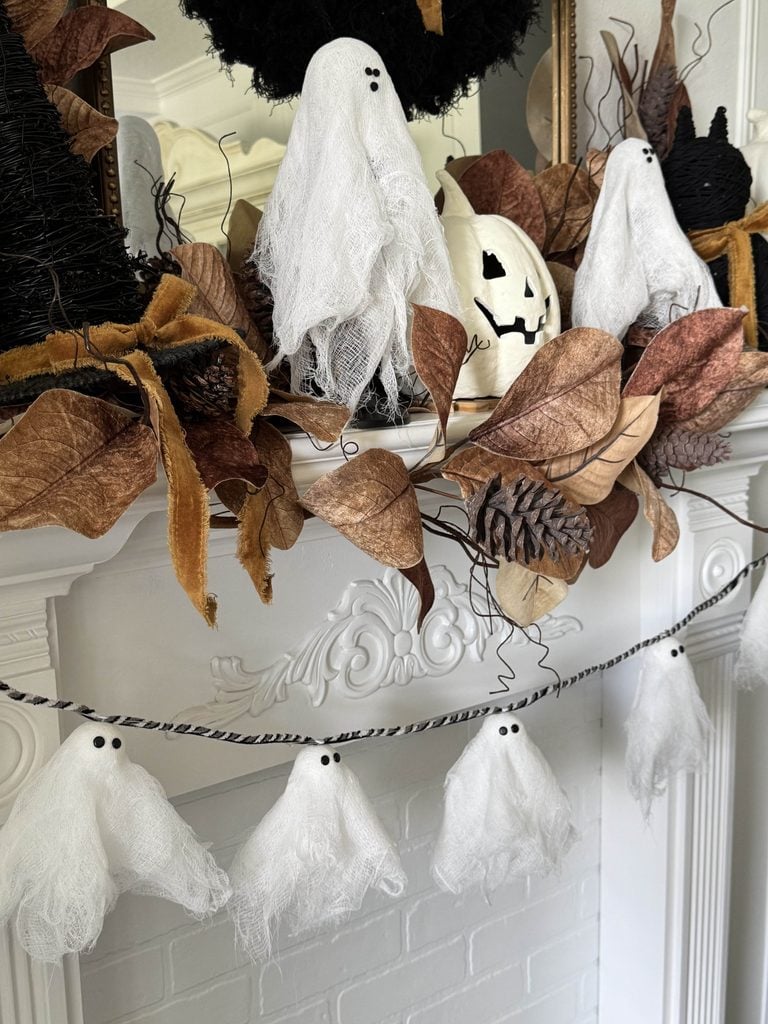 White gauze ghosts and brown leaves decorate a white mantel. A jack-o-lantern, black witch hats, pinecones, and a DIY ghost garland hangs below, creating a festive Halloween scene.