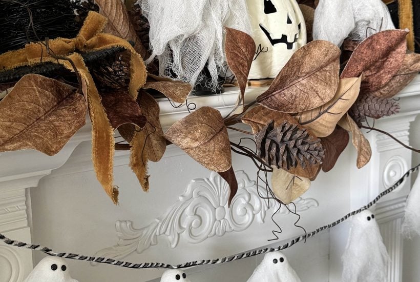 White gauze ghosts and brown leaves decorate a white mantel. A jack-o-lantern, black witch hats, pinecones, and a garland with mini ghosts hang below, creating a festive Halloween scene.