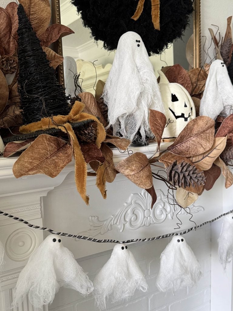A white mantle decorated with fabric ghosts, dried brown leaves, a black witch hat, a white pumpkin, and ghost garland.