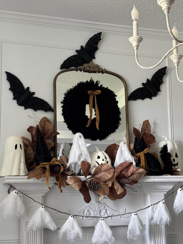 A Halloween mantel decor scene featuring ghost and bat decorations, a black wreath with a gold ribbon, faux leaves, pumpkins, and a garland of white ghost shapes hanging below.