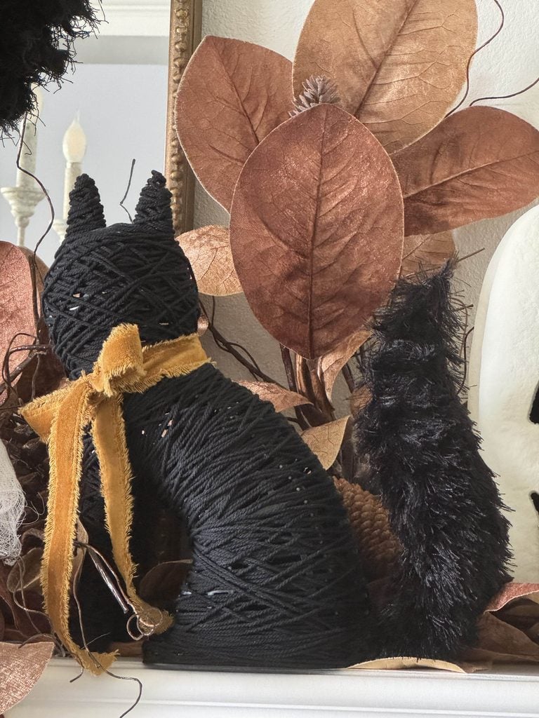 A black yarn-wrapped cat with a gold scarf sits among brown artificial leaves, a fluffy black tail, and a white pumpkin.
