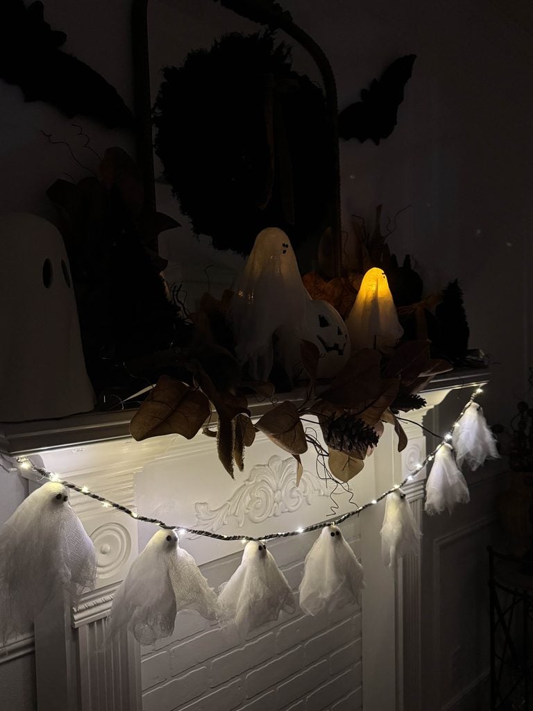 A fireplace mantel decorated with ghost figures, leaves, and a DIY ghost garland with lights, creating a spooky Halloween atmosphere. A small candle-lit ghost glows among the decorations.
