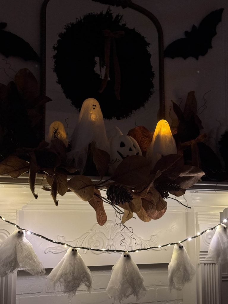 A Halloween mantel decor setup with glowing ghost figures, a white pumpkin, dried leaves, pinecones, a black wreath, and string ghost ornaments. A black bat silhouette completes the spooky look.