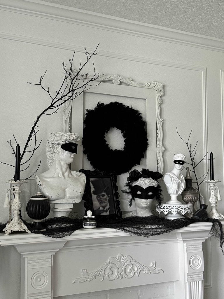 A Halloween mantel decor featuring white busts with black masks, black candles, bare branches, a black wreath, and a framed black-and-white photo creates an elegant, gothic, and mysterious atmosphere.