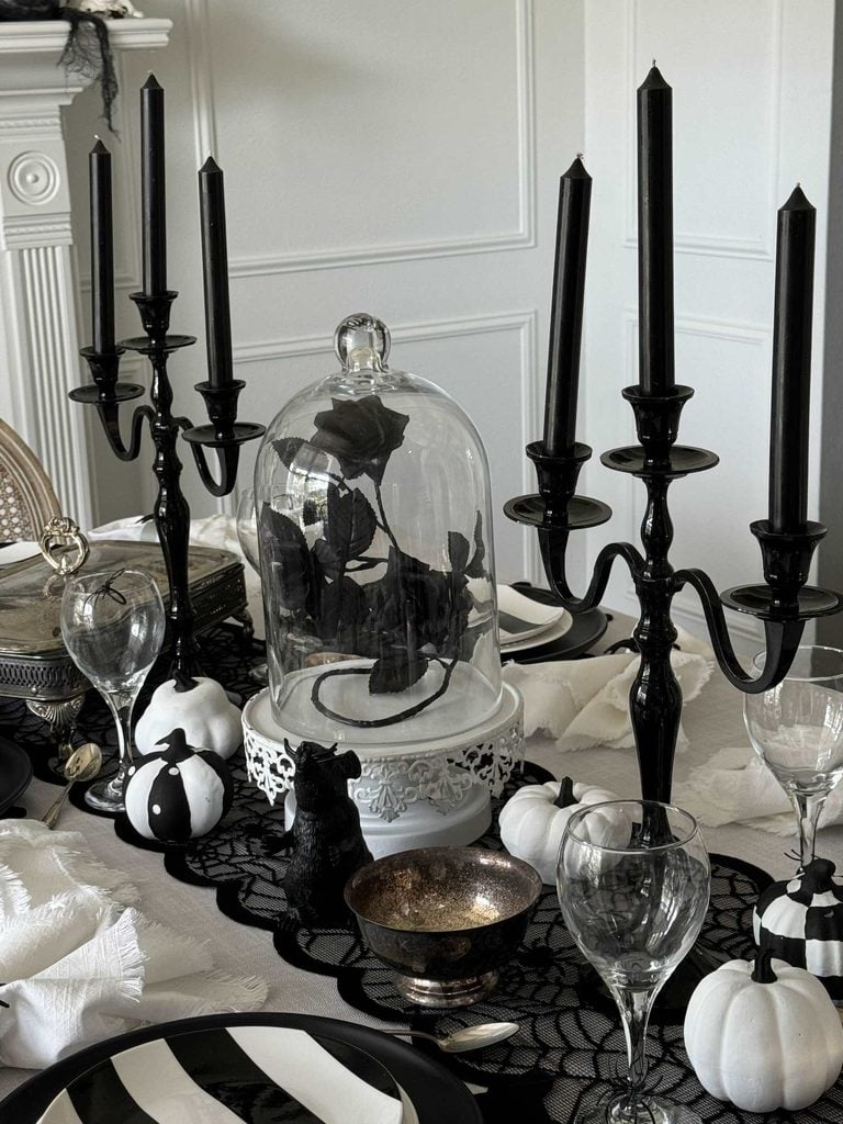Elegant Halloween table setting with black candles, glass cloche covering black roses, and black and white pumpkins evokes chic Halloween mantel decor atop a lace tablecloth. Glassware and napkins complete the monochrome gothic look.